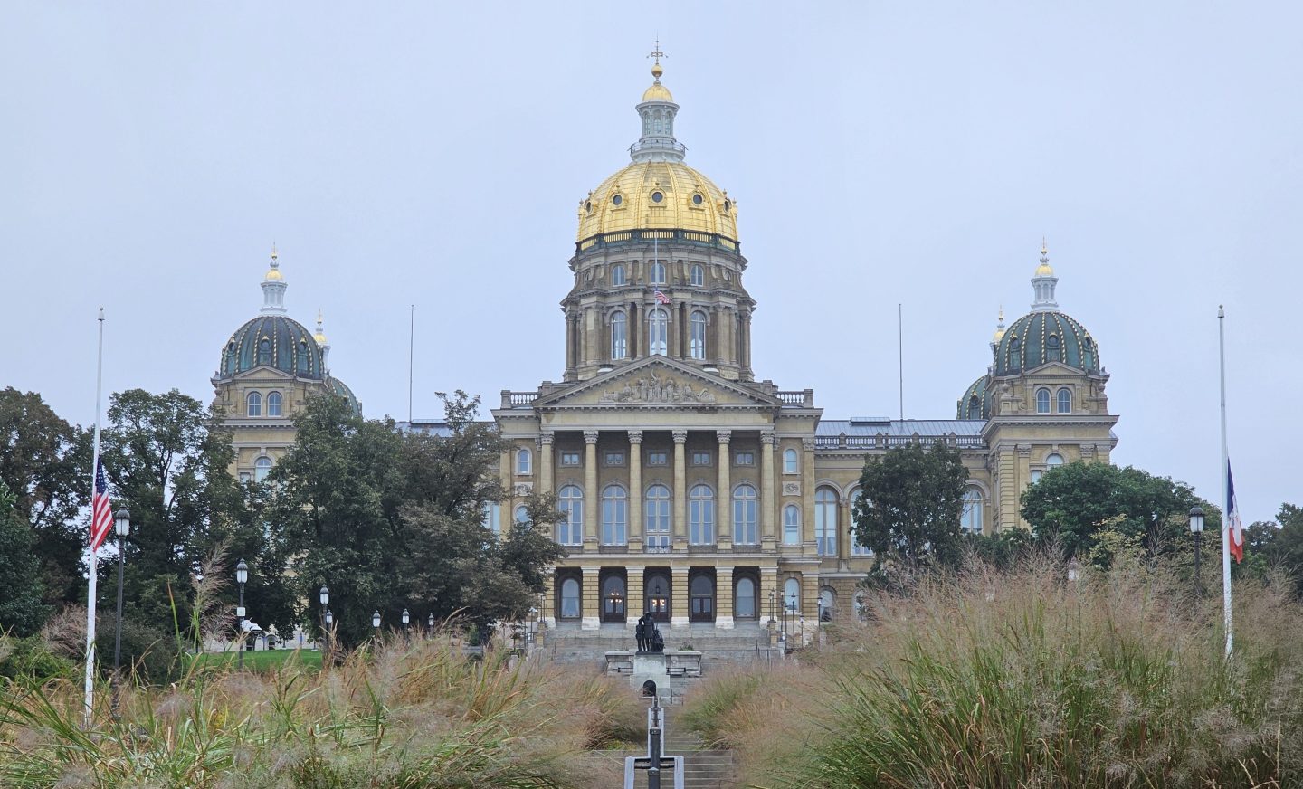 Iowa State Capitol building with a golden dome, view from the front surrounded by greenery and flags.